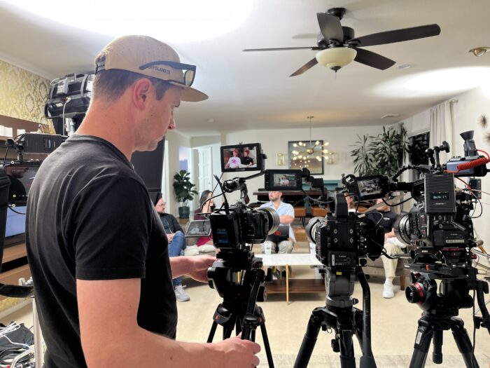 Three cinema cameras and a videographer are set up to record, with subjects on a couch in the backgroun